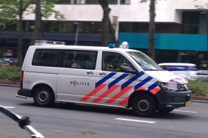 Ein Polizeivan fährt auf einer Straße neben einem Fahrrad, mit Menschen im Van und einem Gebäude, Bäumen und Pflanzen im Hintergrund.