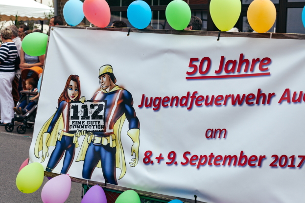 Eine Gruppe von Menschen auf einer Straße, einige mit Kinderwagen, mit einem Banner "50 Jahre Jugendfeuerwehr August 2017" mit Text- und Cartoonsymbolen, einem Zaun, Luftballons, einem Zelt, einem Gebäude mit Fenstern und Bäumen im Hintergrund.