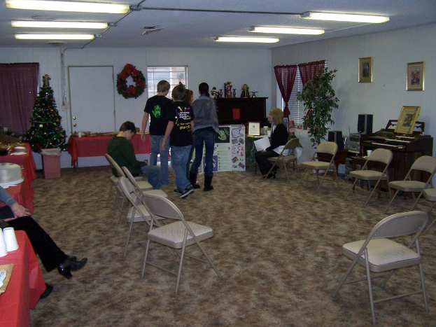 Ein Raum mit mehreren St├╝hlen, Tischen und Menschen, einige stehen und einige sitzen, mit Tassen und Gef├Ą├čen auf den Tischen, einer Wand im Hintergrund mit einem Strau├č, Vorhang, T├╝r, Fotorahmen und Fenstern, einem Weihnachtsbaum, einer Orgel auf der rechten Seite und Röhrenleuchten an der Decke.