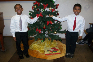 Ein geschmückter Weihnachtsbaum steht in einem Raum mit einem Holztisch links und einem Stuhl rechts, zwei Jungen in weißen Hemden lächeln und halten den Baum, eine Schachtel und andere Gegenstände auf dem Boden und eine weiße Wand mit angebrachten Gegenständen im Hintergrund.