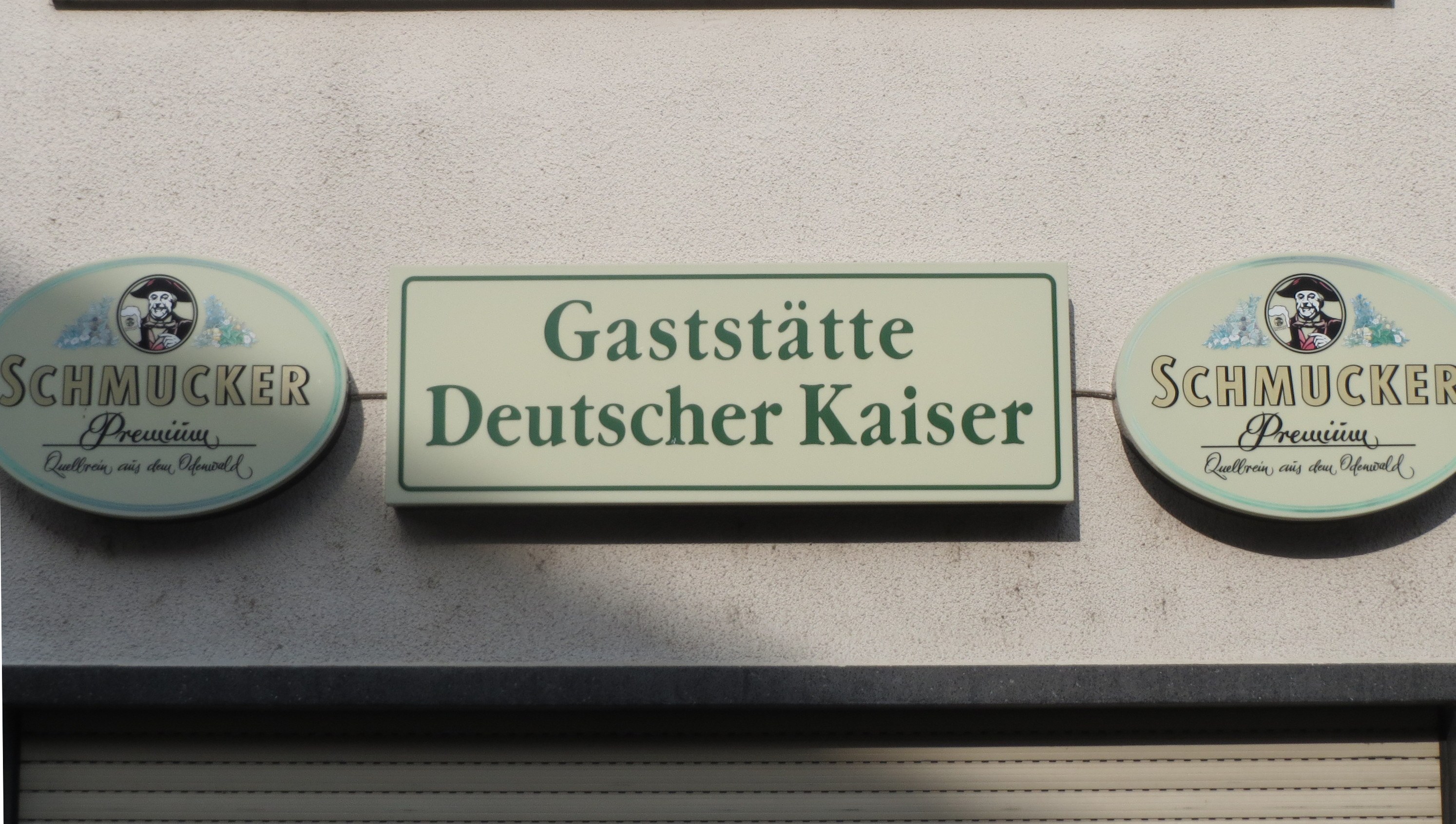 Drei hölzerne Bretter mit der Aufschrift "Gaststätte Deutscher Kaiser Schmucker" sind an einer Gebäudewand über einem geschlossenen Rollladen angebracht.