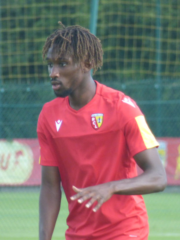 Ein Mann mit Dreadlocks steht auf einem Fussballfeld und trägt ein rotes T-Shirt und Shorts, im Hintergrund gibt es ein Netz und eine Fahne mit der Aufschrift "RCCL - Ghanaian Premier League"