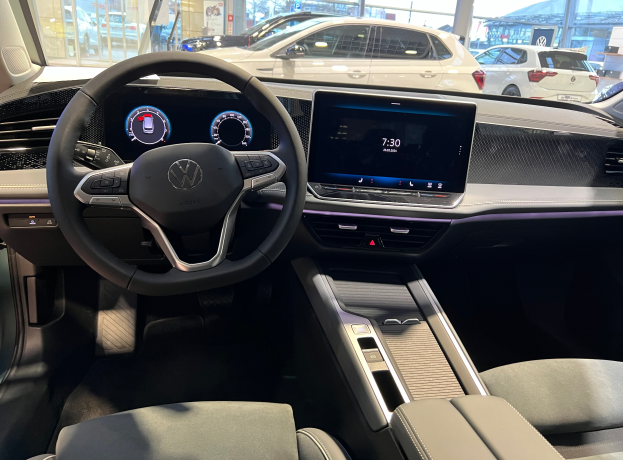 Das Innere eines Volkswagen e-Golf mit einem Lenkrad, Armaturenbrett, Bildschirmen, Knöpfen, Sitzen und Glastüren, mit Blick auf andere Autos, Gebäude, Bäume, Pfähle und den Himmel.