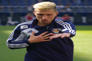 Ein junger Mann in einem blauen T-Shirt steht auf einem Fußballfeld und streckt die Arme aus, wobei er entschlossen wirkt und der Hintergrund unscharf ist.