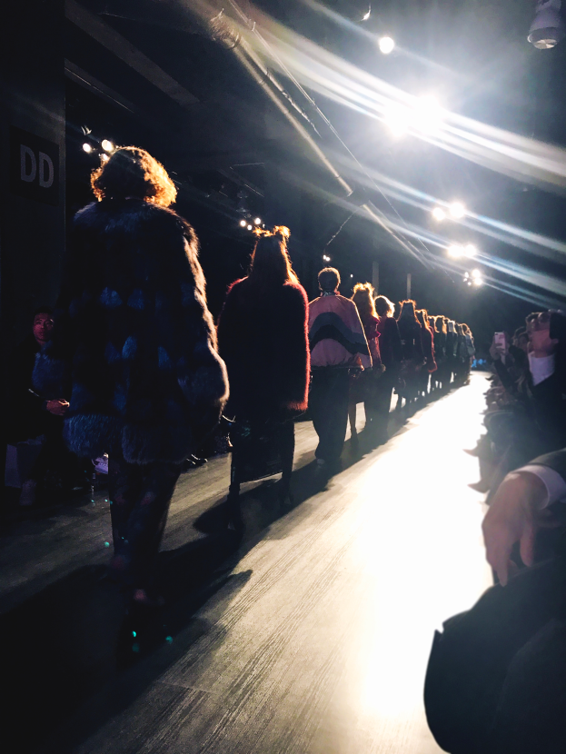 Eine Gruppe von Menschen läuft über einen Laufsteg bei einer Modenschau, während sie von hellen Scheinwerfern beleuchtet wird und Zuschauer auf beiden Seiten zuschauen.