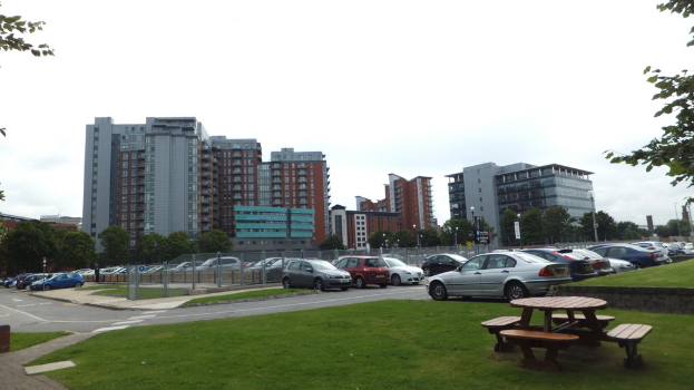 Ein Parkplatz mit vielen Autos, umgeben von Gras, Bäumen, Gebäuden und einem klaren Himmel, mit einer Bank mit Stühlen auf dem Rasen.