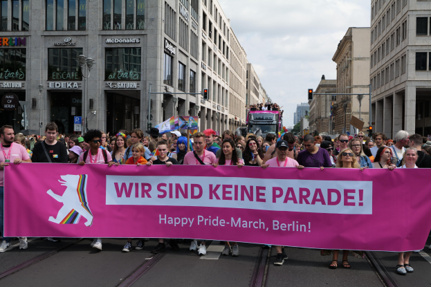 Eine Gruppe von Menschen marschiert auf einer Straße in Berlin mit einer pinken "Happy Pride March"-Fahne, umgeben von Gebäuden, Laternenmasten und Verkehrszeichen unter einem bewölkten Himmel.