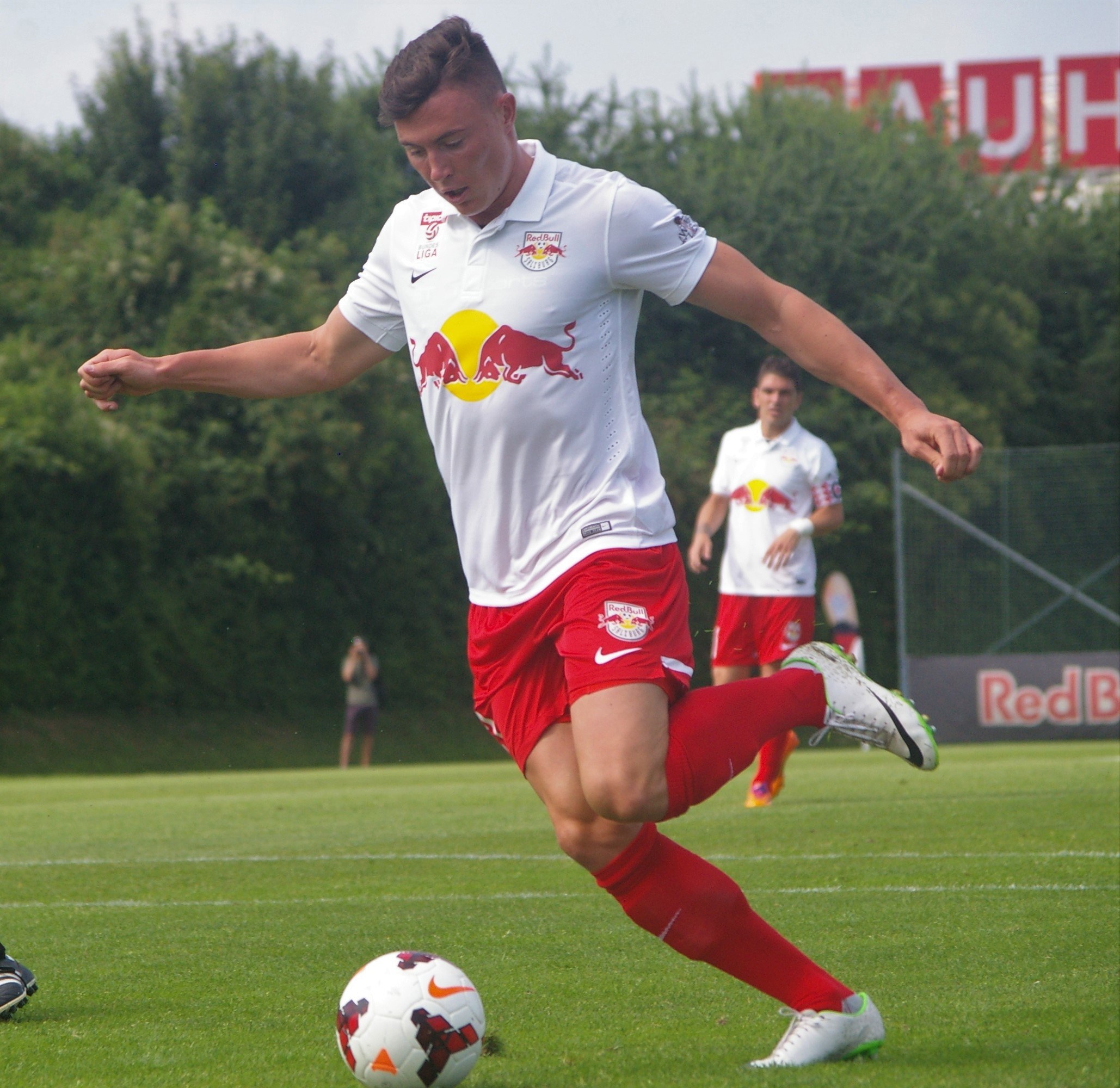 Ein professioneller Fußballspieler, der einen Fußball auf einem Rasenfeld kickt, mit "RB Leipzig" im Hintergrund bei klarem blauem Himmel.