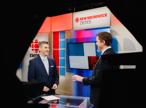 Zwei Männer in Blazern und Krawatten vor einem Glastisch mit Papieren, mit einem Fernseher im Hintergrund, der 'New Brunswick News' zeigt.