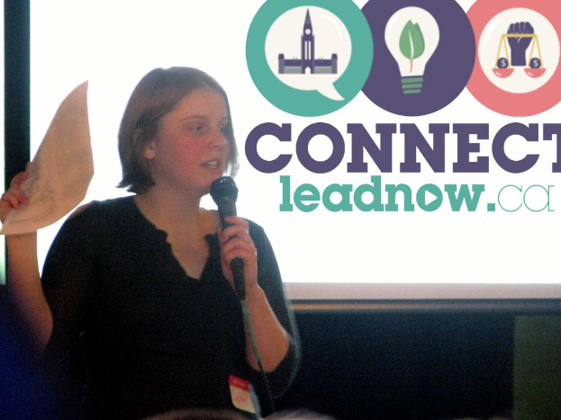 Eine Frau hält ein Mikrofon und Papier und spricht, während im Hintergrund ein Bildschirm mit Text und Logos zu sehen ist.