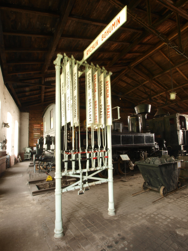 Ein Bahnsteig mit einem Zug auf den Gleisen, ein Bodenwagen, ein Signalhaus in der Mitte, ein Pfahl mit einer angebrachten Tafel, eine Wand links und eine Decke darüber.