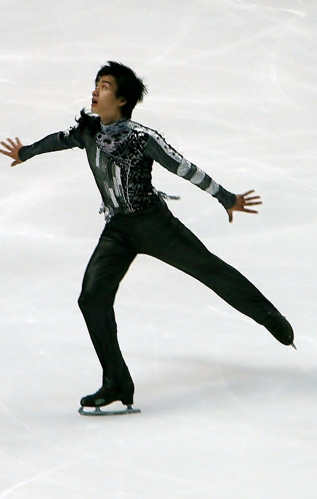 Männlicher Eiskunstläufer in einem schwarzen Kleid, der eine Routine auf einer Eisfläche zeigt, gleitet mit synchronisierten Arm- und Beinbewegungen.