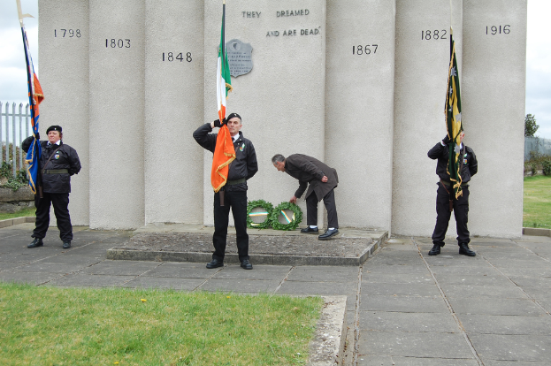 Drei Menschen in Uniform stehen mit Fahnen verschiedener Farben, dahinter ein Mann mit grÃ¼nen KrÃ¤nzen, vor einer Wand oder SÃ¤ulen mit Text, Gras, Gehweg, einem Zaun auf der linken Seite und BÃ¤umen und einem GebÃ¤ude auf der rechten Seite.