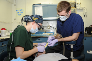 Zwei Chirurgen in Masken und Handschuhen operieren einen Patienten in einem Zahnarztstuhl, mit verschiedenen Gegenständen auf dem Tisch und Rohren und einer Tafel im Hintergrund.