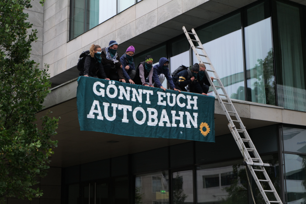 Eine Gruppe von Menschen steht auf dem Dach eines Gebäudes und hält ein Transparent in die Höhe, links neben ihnen ein Baum und im Hintergrund Glasfenster, sie scheinen für die deutsche Regierung zu protestieren.