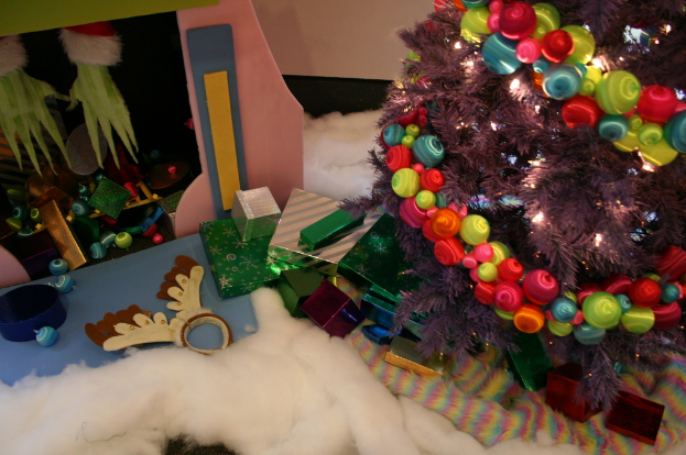 Drinnen steht rechts ein Weihnachtsbaum mit Lichterkette und Schmuck, umgeben von Geschenkboxen und Dekorationsgegenständen, mit einer Hütte und Watte oben links und einem Wolltuch unten.