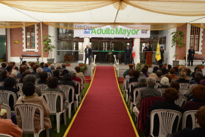 Ein Mann steht vor einem Geb├Ąude mit einem Bogen, auf dem "Adulto Mayor" steht, w├Ąhrend viele Menschen im Hintergrund auf St├╝hlen sitzen.