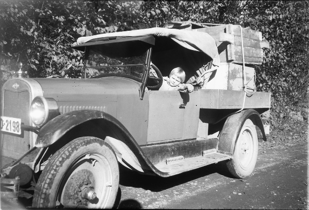Altes Schwarz-Weiß-Foto eines Mannes und Kindes, die gemeinsam auf dem Fahrersitz eines Lastwagens auf einer Straße mit Bäumen im Hintergrund sitzen.