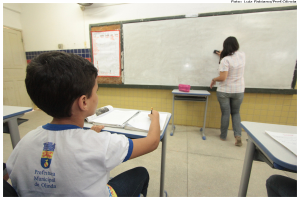 Ein Junge sitzt an einem Tisch und schreibt in ein Buch, während eine Frau ein Brett mit einem Putzlappen in einem Klassenzimmer mit mehreren Tischen, einem mit einer Box und einem Plakat an der Seitenwand putzt.