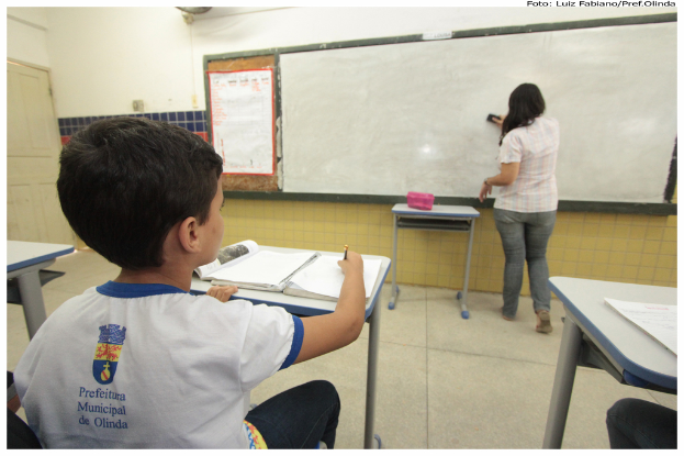 Ein Junge sitzt an einem Tisch und schreibt in ein Buch, während eine Frau ein Brett mit einem Putzlappen in einem Klassenzimmer mit mehreren Tischen, einem mit einer Box und einem Plakat an der Seitenwand putzt.