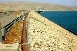 Ein großes Rohr liegt auf der Seite einer Straße neben einem Gewässer, mit einer Brücke im Hintergrund, fernen Hügeln und einem klaren blauen Himmel, umgeben von Steinen und Gras, mit Text am unteren Bildrand.