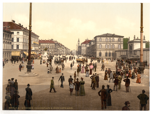 Ein altes Foto einer belebten Stadtstraße in München, Deutschland, aus den frühen 1900er Jahren, mit Passanten, einige mit Schirmen, einem Pferdewagen im Vordergrund, Gebäuden, Bäumen und Polen im Hintergrund, mit Text am unteren Bildrand.