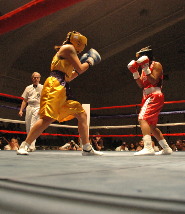 Zwei Boxer kämpfen im Ring mit einem Schiedsrichter im Hintergrund, umgeben von Fotografen und Zuschauern unter einer sichtbaren Decke.