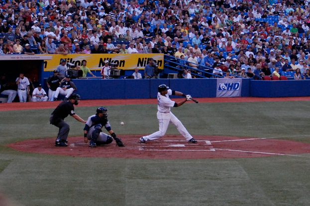 Ein Mann hält einen Baseballschläger mit einem Ball hinter sich, während mehrere Personen auf Stühlen sitzen und andere Kameras in der Hintergund halten, mit Werbetafeln und Metallstäben davor.