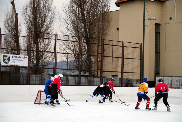 Menschen spielen Eishockey auf einem Eis mit Gebäuden, Bäumen, einer Straßenlaterne, einem Namensschild und Zäunen im Hintergrund unter einem Himmel.