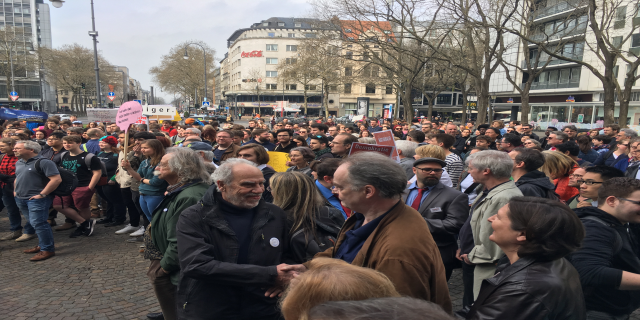 Eine große Gruppe von Menschen, die vor einer Menge mit Schildern steht, mit Fahrzeugen, Bäumen, Laternenmasten und Gebäuden im Hintergrund, wahrscheinlich während einer Demonstration in Berlin, Deutschland.