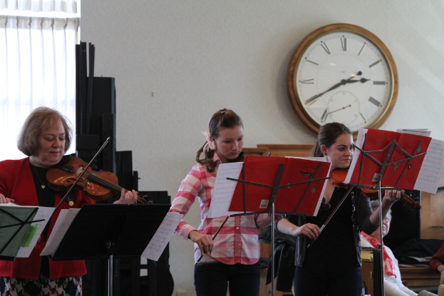 Drei Frauen auf der Bühne mit Violinen stehend und spielend, mit Notenpulten und Noten davor, während ein Zuschauer im Vordergrund sitzt und zuhört; eine Uhr hängt an der weißen Wand im Hintergrund, und ein weißer Vorhang ist links sichtbar.