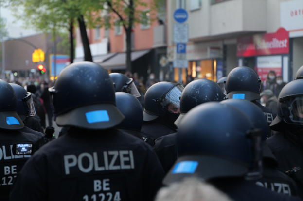 Eine Gruppe von Polizisten in schwarzen Uniformen und Helmen steht vor einer unscharfen Menge und einer Stadtlandschaft mit Gebäuden, Bäumen, Polen, Verkehrszeichen und Schildern.