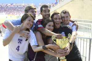 Sechs Menschen, zwei Frauen und vier Männer, halten einen Pokal und lächeln für ein Foto vor einem Eisengeländer, mit einer Wand und einem Geländer oben rechts, Menschen im Hintergrund und sichtbare Lichter, wahrscheinlich in einem Stadion.