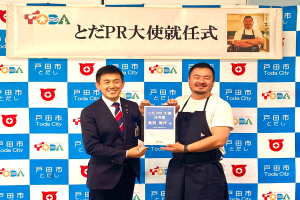 Zwei lächelnde Männer mit einem Zertifikat, mit einem Banner im Hintergrund, das "Toda City Wins PR Award" lautet und ein Foto einer Person zeigt.