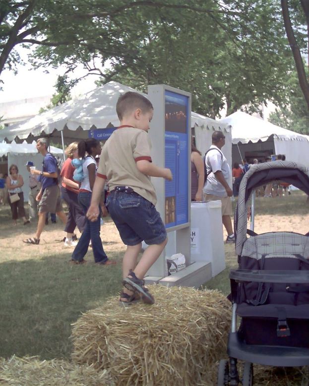 Eine Szene mit mehreren Zelten und Menschen, die darum herum gehen, ein Displaybrett vorne, ein Kind, das auf trockenem Gras daneben spielt, und Bäume auf beiden Seiten.