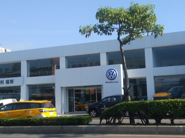 Volkswagen-Händler mit Glaswänden, parkenden Autos, umgeben von Pflanzen und einem Baum.