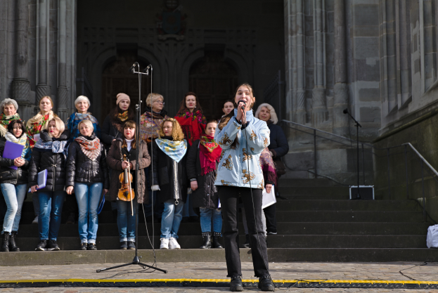 Eine Gruppe von Menschen, einige mit Musikinstrumenten, steht vor einem Gebäude mit Stufen und Geländern, während eine Frau in der Mitte in ein Mikrofon singt, umgeben von zusätzlichen Mikrofonen auf Ständern.