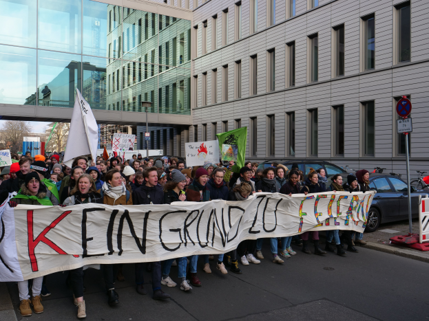Eine Gruppe von Menschen steht auf einer Straße und hält ein Transparent mit der Aufschrift "Kein Grind Lo Feuer" im Protest, mit Gebäuden, Bäumen, Polen, Schildern, Fahrzeugen und einem klaren blauen Himmel im Hintergrund.