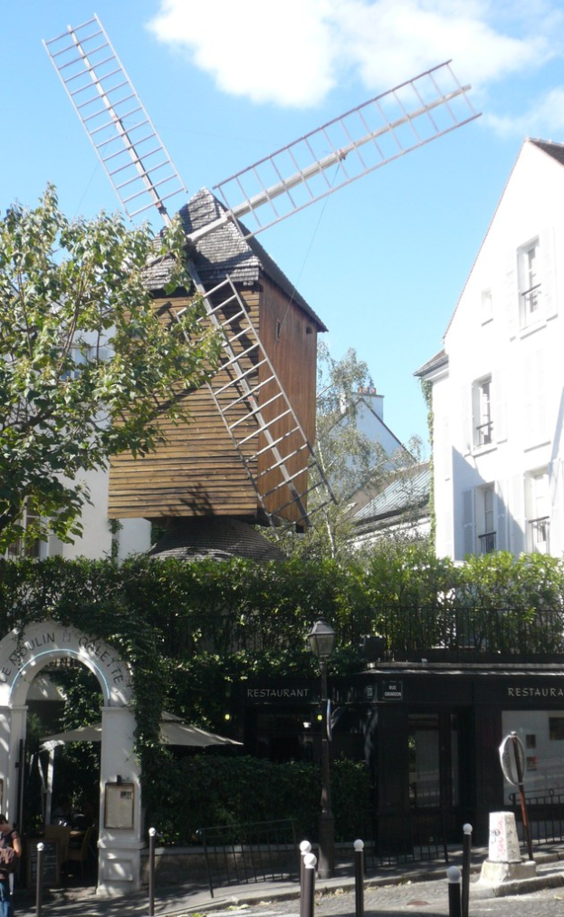 Eine Windmuehle, mehrere Gebaeude, Baeume, Metallstangen und ein Zaun in einer landlichen Umgebung.