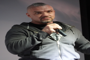 Komiker in einem schwarzen Anzug und einem wei├čem Hemd, der ein Mikrofon h├Ąlt und selbstbewusst vor einer Wand steht.