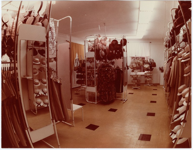 Altes Foto eines Kleiderladens mit Kleidung an der Stange, Hüten auf Ständern, einem Spiegel, einem Stuhl, einem Tisch mit Gegenständen, Vorhängen und Deckenleuchten.