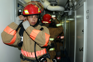 Eine Gruppe von Feuerwehrleuten in Uniform steht zusammen in einem Raum mit einer Tür auf der rechten Seite und einer Wand auf der linken Seite, mit Rohren und anderen Gegenständen im Hintergrund.