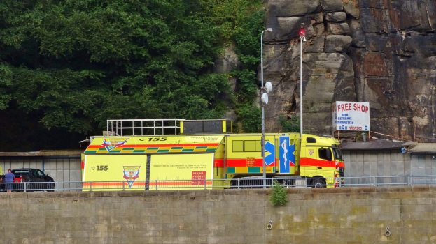 Ambulanz auf der Seite einer Straße neben einer Klippe geparkt, mit einer Wand, Gelönder, Pfosten, Lichtern, Schildern mit Text und anderen Gegenständen im Vordergrund und Bäumen und einem Auto im Hintergrund.