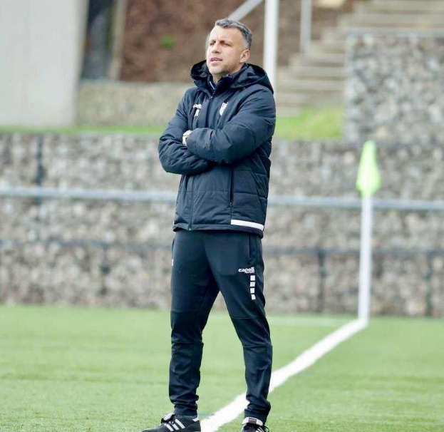 Ein Füßballtrainer, der auf einem Feld steht und die Ärmel überkreuzt, trägt eine schwarze Jacke und Hose, mit einem Zaun, Treppe und Wand im Hintergrund.