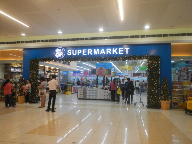 Menschen vor einem Supermarkt mit Einkaufstaschen, einem Einkaufswagen, einem Topf mit Pflanze, Regalen, Schildern und Deckenbeleuchtung.