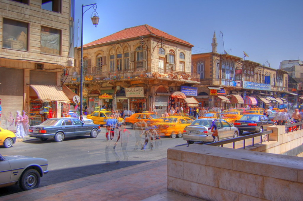 Menschen gehen auf einem Gehweg, w├Ąhrend Autos auf der benachbarten Stra├če fahren, mit Geb├Ąuden, Laternenmasten und Gesch├Ąften ├╝ber die Stra├če hinweg sichtbar.