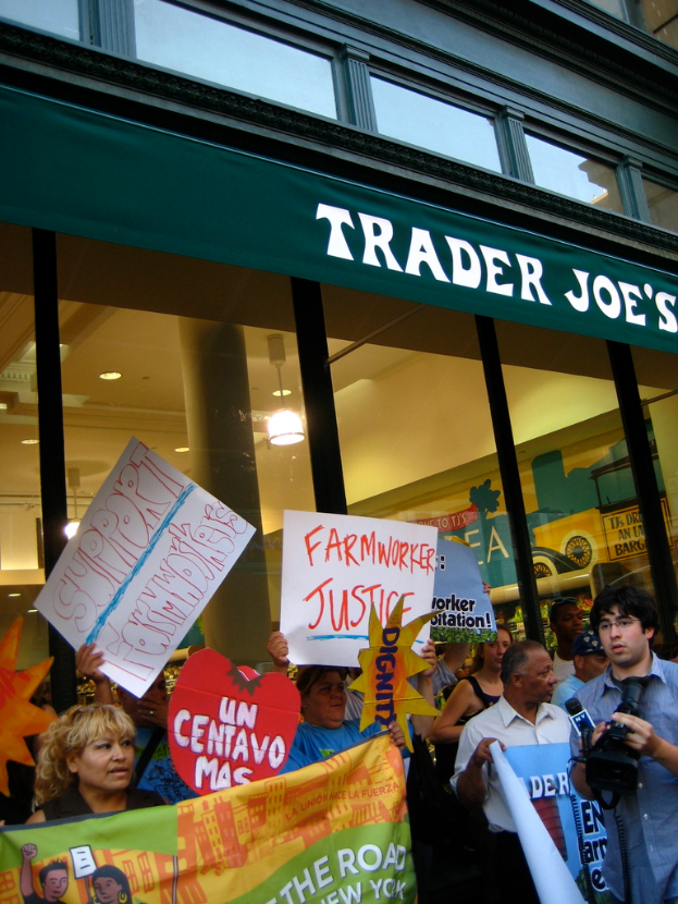 Eine Gruppe von Menschen steht vor einem Geschäft und hält Schilder und Plakate hoch, mit einer Säule und Lichtern auf dem Dach im Hintergrund.