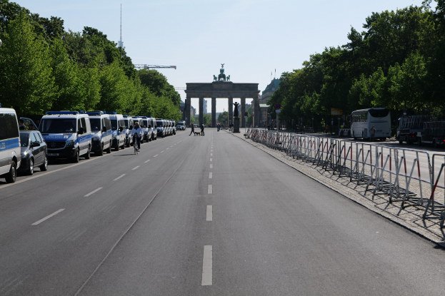 Lange Reihe von Polizeiwagen, die an einer Straße vor dem Brandenburger Tor geparkt sind, mit Fahrradfahrern, Fußgängern, Barrieren und Bäumen; ein prunkvoller Bogen mit Statuen im Hintergrund unter einem sichtbaren Himmel.