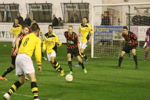 Ein Sportplatz mit mehreren Männern, die Fußball spielen, ein Netz auf der rechten Seite, eine Tafel mit Text im Hintergrund, Zuschauern hinter der Tafel und einer Wand mit Fenstern im Hintergrund.