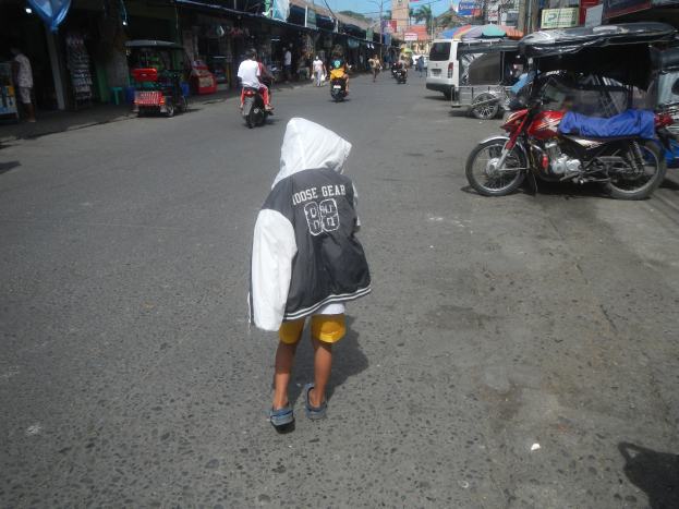 Ein junger Junge geht eine Straße entlang, die von Motorrädern und anderen Fahrzeugen bevölkert ist, mit Gebäuden, Bäumen und einem klaren blauen Himmel im Hintergrund.